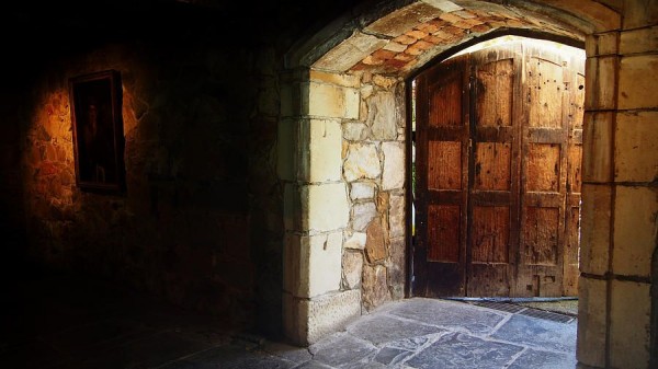 A photograph of a dungeon door, partially open to outdoors. A portrait is in the darkness to its left. By Lois Romer.